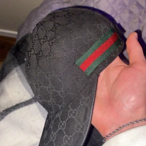 Svart Gucci keps med GG-mönster - Säljer en svart keps från Gucci med klassiskt GG-monogram över hela kepsen. Sidan har den ikoniska gröna och röda randen. Justerbar rem baktill och insida med Gucci-tag. Perfekt för dig som vill ha en lyxig och stilren accessoar.