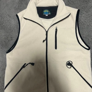 Beige fleeceväst från True North - Beige fleeceväst från True North med svarta detaljer och dragkedjor. Västen har hög krage, tre fickor med dragkedja och svart kantband runt ärmhål och dragkedja. Perfekt för lager-på-lager och outdoor vibes. Den är strl S men passar en större M och L. 