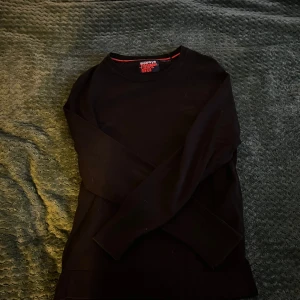 Svart tröja från Superdry Orange Label - Svart långärmad tröja från Superdry Orange Label med rund halsringning och diskret röd detalj på insidan av kragen. Klassisk och enkel design i mjuk bomull, perfekt för en clean och stilren look.