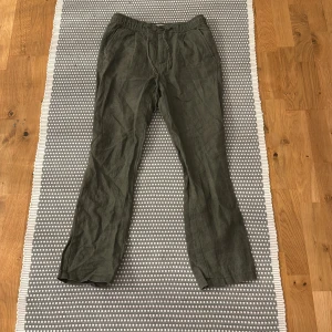 Olivgröna linnebyxor Knowledge Cotton - Snygga olivgröna linnebyxor från Knowledge Cotton Apparel med elastisk midja och raka ben. Byxorna har fickor både fram och bak samt knappstängning framtill. Perfekta för en chill och avslappnad stil under varmare dagar.