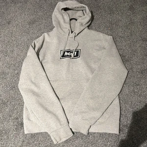 Grå Hov1 hoodie med broderad logga - Säljer en grå hoodie från Hov1 med broderad svartvit logga på bröstet. Tröjan har huva med snörning och en klassisk känguruficka framtill. Perfekt för dig som gillar streetwear och vill ha något bekvämt och stilrent.