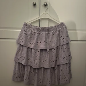 Ljuslila volangkjol från Vila - Supersöt ljuslila kjol från Vila med flera volanger och broderade hålmönster. Kjolen har resår i midjan och är i storlek XS/S. Kjolen slutar strax ovanför knäna.
