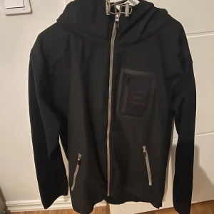 Svart Hugo boss  - Svart vindjacka från Boss med grå dragkedjor, huva och praktiska fickor framtill. Jackan har en bröstficka med Boss-logga och justerbar dragsko vid huvan. Perfekt för blåsiga dagar och snygg till streetwear.