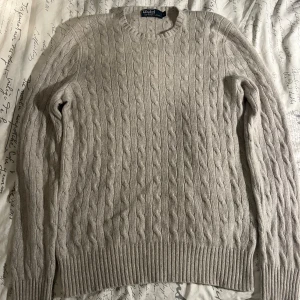 Beige kabelstickad kashmirtröja Polo - Säljer en beige kabelstickad tröja från Polo Ralph Lauren i 100% kashmir. Tröjan har rund halsringning, ribbade muddar och lång ärm. Perfekt för dig som vill ha en lyxig och mjuk tröja med klassisk look.