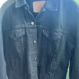 Svart jeansjacka från Levi's, M - Säljer en svart jeansjacka från Levi's i klassisk modell med knappar framtill och två bröstfickor med lock. Jackan har en snygg, tidlös look och är tillverkad i bomull.  ikonisk denimjacka ! Storlek M  ca 50-52 .