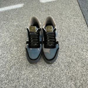 Valentino Garavani Rockrunners - Snygga Valentino Garavani sneakers med blått och svart camouflage-mönster, svarta snören och beige detaljer. Ovandelen är i mesh och skinn, med en robust gummisula och rund tå. Perfekt för dig som gillar exklusiv stil med sköna vibbar.