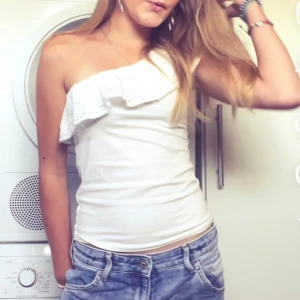 Vit oneshoulder topp Holly & Whyte - Supersnygg vit oneshoulder topp från Holly & Whyte by Lindex i storlek S. Toppen har en bred volang längs halsringningen och är figurnära med axelbandslös design. Perfekt för sommarens alla tillfällen, matcha med jeans eller kjol för en fräsch look.