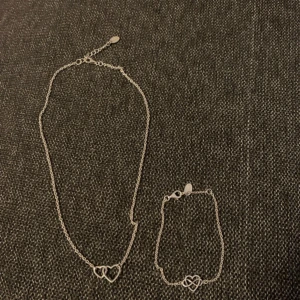 Sahara halsband och armband i silver - Set med halsband och armband från Sahara i silverfärgad metall. Båda har tunna kedjor och fina hjärtformade detaljer, där halsbandet har två sammanlänkade hjärtan och armbandet ett hjärta med oändlighetssymbol. Perfekt för dig som gillar stilrena smycken med en romantisk touch.
