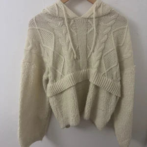 Beige stickad hoodie med flätmönster - Supermysig beige hoodie med stickat flätmönster och ribbade detaljer. Tröjan har huva med snörning och är tillverkad i en mix av stickat och teddy-liknande material på ärmar och nedre delen. Perfekt för kalla dagar och chill vibes.