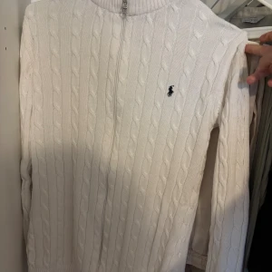 Vit kabelstickad tröja från Ralph Lauren - Kabelstickad vit tröja från Ralph Lauren med halv dragkedja och klassisk broderad logga på bröstet. Tröjan har lång ärm, ribbade muddar och hög krage. Perfekt för en clean och stilren look. Storlek är ack men jag hade sagt att den är mer L