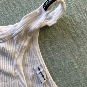 Vit ribbad tanktop från Dolce & Gabbana - Säljer en vit ribbad tanktop från Dolce & Gabbana. Toppen har klassisk rund halsringning och ärmlös design med breda axelband. Materialet är mjukt och stretchigt, perfekt för en clean och stilren look. Passar dig som gillar exklusiva märken och enkel stil.
