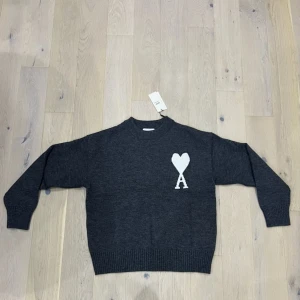 Mörkgrå stickad tröja från Ami Paris - Mörkgrå stickad tröja från Ami Paris med vit hjärtlogga och bokstaven A på bröstet. Tröjan har rund hals, ribbade muddar och är långärmad. Perfekt för dig som gillar stilrena och trendiga plagg med en twist.