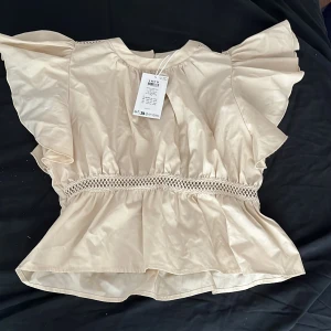 Beige blus med volang från LMTD - Supersöt beige blus från LMTD med volangärmar och markerad midja. Blusen har dekorativ spetsdetalj runt midjan och knäppning med pärlknapp i nacken. Tillverkad i ett glansigt, lätt material som ger en lyxig känsla. Tröjan har aldrig används.