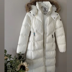 Vit lång dunjacka från Moncler - Snygg lång vit dunjacka från Moncler med avtagbar huva och lyxig pälskant. Jackan har markerad midja med bredt bälte där Moncler-loggan är broderad, dragkedja framtill och flera fickor med dragkedja, jackan är i nyskick då jag endast haft den 1 kort tillfälle men kände den blev förstor för mig , jackan är i strl 0 som motsvarar en xs men denna är större i strl så skulle säga en small kan ha denna även en liten M om man inte har en jätte stor byst  