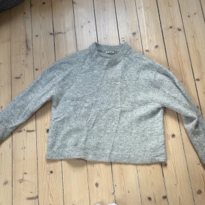 Grå stickad tröja från Gina Tricot - Mysig grå stickad tröja från Gina Tricot med rund halsringning och ribbade muddar. Perfekt för lager-på-lager och har en klassisk, enkel design som funkar till allt. Lätt croppad passform och lång ärm.