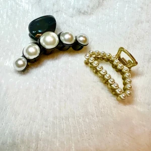 Två små hårklämmor med pärldetaljer - Säljer två snygga hårklämmor, en svart med stora vita pärlor och en guldfärgad med små pärlor runt hela klämman. Perfekta accessoarer för att lyfta din frisyr med en elegant touch.