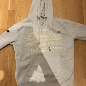 Ljusgrå hoodie från Superdry - Ljusgrå hoodie från Superdry med broderad logga och japanska tecken på bröstet. Tröjan har huva med snörning, känguruficka och ribbade muddar. Perfekt för chill dagar och snygg streetstyle.
