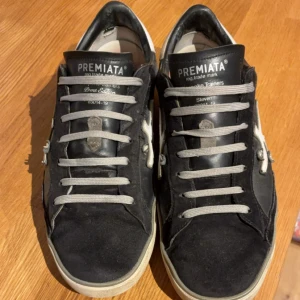 Premiata svarta sneakers med vit detalj - Svarta sneakers från Premiata med vit logodetalj på sidan och vit häl. Skorna har grå snörning och en platt sula med tryck. Materialet är en mix av skinn och mocka, vilket ger en schysst kontrast. Perfekt för dig som gillar stilrena och coola sneakers.