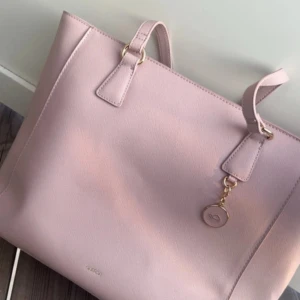 Ljusrosa axelväska från Adax - Säljer en stilren ljusrosa axelväska från Adax i syntetmaterial. Väskan har guldiga metalldetaljer, en dekorativ berlock och dragkedja upptill. Invändigt finns flera fack och en innerficka med dragkedja. Perfekt för dig som vill ha en snygg och rymlig väska.