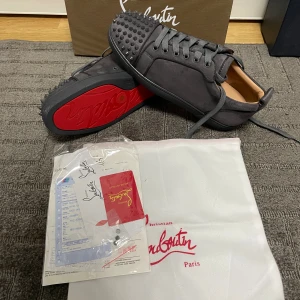 Christian Louboutin gråa sneakers - Snygga svarta sneakers från Christian Louboutin med ikonisk röd sula och coola nitar på tån. Skorna har snörning och är tillverkade i mocka för en lyxig känsla. Perfekta för dig som vill sticka ut med exklusiv streetstyle.
