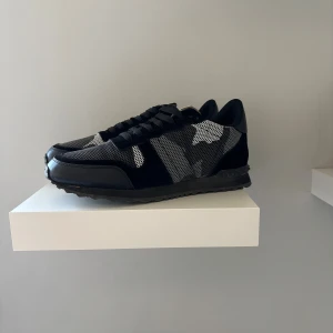 Valentino Garavani Rockrunner sneakers - Snygga Valentino Garavani Rockrunner sneakers i svart, grått och vitt med coolt camouflage-mönster. Skorna har ovandel i mocka och mesh, svarta snören och en robust gummisula. Perfekta för dig som vill sticka ut med exklusiva och trendiga sneakers.