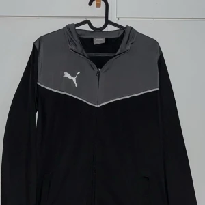 Puma - Kom med pris. Svart och grå hoodie från Puma med dragkedja framtill och huva. Hoodie har en sportig look med kontrastpanel upptill och Puma-logga på bröstet. Perfekt för dig som gillar streetwear och vill ha en lätt hoodie till vardagen.