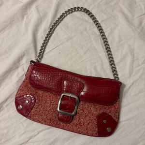 Röd axelväska från Frangi med kedja - Cool röd axelväska från Frangi med grov silverkedja som axelrem. Väskan har detaljer i röd croco-präglad skinnimitation, stor silverspänne framtill och vävd textil i rött och rosa. Perfekt för dig som vill sticka ut med accessoarer.