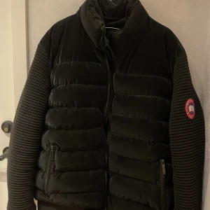 Svart pufferjacka från Canada Goose - Säljer en svart pufferjacka från Canada Goose med stickade ärmar och klassisk logga på armen. Jackan har dragkedja framtill och två fickor med dragkedja. Perfekt för kyliga dagar och har en riktigt clean look.