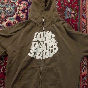 Brun hoodie från Lowlights Studios M - Brun hoodie från Lowlights Studios i storlek medium med stor vit grafisk logga på framsidan. Tröjan har huva, dragkedja och två fickor framtill. Mjuk insida och klassisk streetwear-look, perfekt för chill dagar.