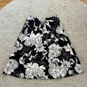 Svartvit blommig kjol från H&M - Svart A-linjeformad kjol från H&M med stora vita blommor. Kjolen är knälång och har dragkedja i sidan. 