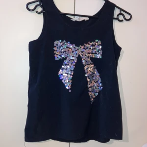 Svart festtopp med paljetter  - Säljer detta snygga linnet med paljetter format som en rosett på, från h&m, då den blivit för liten. Använd fåtal gånger och är i bra skick! 🎀