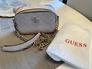 Guess väska  - Liten axelväska i “kamera”-modell. Axelrem/kedja med insättning, dubbla dragkedjor – ena facket med invändig dragkedjeficka, det andra med två kortfack. Mått ca 20*13*7 cm Nypris: 1  045 kr Köpt på Rizzo.   Använd, men i bra skick.  