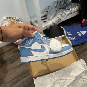 Air Jordan 1 mid (Blue and White) storlek 36 - Helt oanvända Jordan skor pågrund av att den var liten för personen jag köpte den till. Skorna har med sig 2 par sko knuten med ena färgen blå och andra färgen vit. Skorna är också väldigt fina och det finns även kartong.