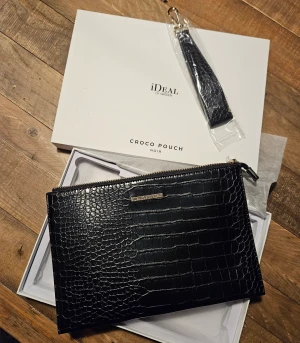 Croco Pouch från Ideal of Sweden - Croco Pouch från Ideal of Sweden är en klassik modell med krokodil-inspirerad textur. Har två fickor och utrymme för 6 kort. Med diskret logga i guld, inkluderar avtagbart band. Storlek 26×18×2 cm. Nypris 700 kr.