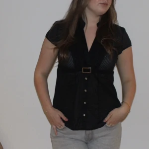 90's vibe top - Beställningar görs på vår hemsida: thrifteddesign.one.    En svart kortärmad top i ett tunt svart tyg med ett silvrigt metallspänne framtill och knappar. 