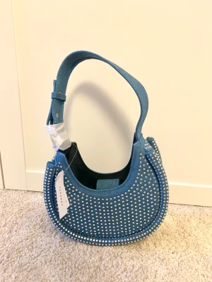Oanvänd väska från ASOS - Helt oanvänd jeansväska med diamanter på🩵 Nypris 369kr. Väskan stängs med hjälp av en knapp och har ett kortfack innuti🤗 Köparen står för frakten💕