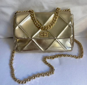 Väska - Guldfärgad väska köpt på primadonna collection i Rom. Endast använd några få gånger och är som i nyskick. Mått: Längd: 30 cm. Höjd: 19 cm. Bredd: 9 cm. 