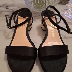 Snygga Sandaletter med guldig klack - Svarta sandaletter i tyg med  spänne storlek 36. Knappt använda. Remmarna är fodrade.Vädigt fina klackar i guldfärg. Bekväma! 