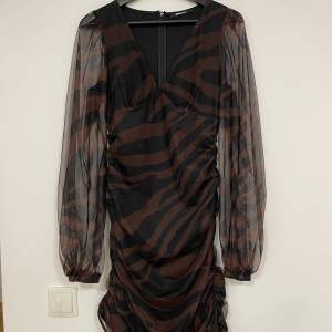 En brun/svart randig klänning från gina i strl S. Aldrig använd. V ringning i fram och genomskinliga ärmar. Jag är 165 cm lång och klänningen går till knäna. 
