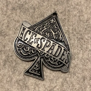 Berlock motörhead - Ace Of Spades, någon slags berlock. Det finns ett litet hål längst upp iallafall. Bild två med bibliotekskort som storleksreferens. Okänt material.