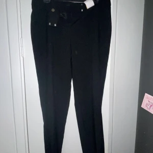 Kostymbyxor svarta - Svarta kostymbyxor i en tajt modell. Helt oanvända, lappen sitter kvar. Både knapp och spänne i midjan för att knäppa byxorna. Vet ej vart dom är ifrån. Står size 14 standard length på lappen, skulle säga att det motsvarar en 38/40 /M