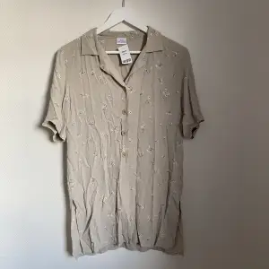 En grågrön skjorta med blom tryck. Skjortans knappar slutar mitt på och nederdelen av skjortan är helt öppen. Den är köpt second hand men jag har sldrig använt den.