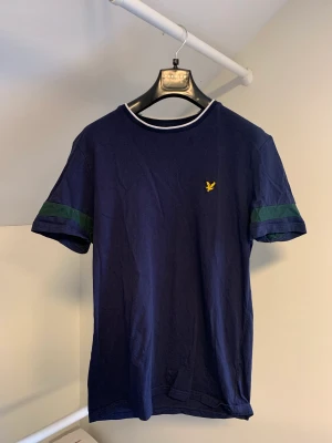 Lyle & Scott t-shirt  - En mörkblå Lyle & Scott t-shirt i storlek S.  Mönster på både kragen och ärmarna där passformen är normal