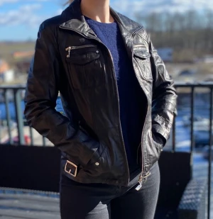 Svart skinnjacka - Klassisk svart skinnjacka från märket Petroff. Storlek (svensk)38. Jackan har innerficka med knapp på vänster sida.  Svart skinnbälte i midjan med silvriga detaljer.  Dubbla bröstfickor på bägge sidor, ena med dragkedja och andra med knapp.