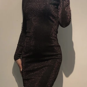 Fest klänning - Glittrig klänning med skönt meterial på insidan. Har en svart basfärg med rosa glitter som skiner i röd/rosa färg. Klänningen är öppen vid övre delen av ryggen. Har stretchigt material och formar sig runt kroppen utan att vara tajt. Aldrig använt.