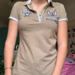 y2k t-shirt - Beige piké t-shirt med mörklblåa och silvriga detaljer. Även text med rhinestones där bak. Super snygg och cyber y2k stil vilket är väligt inne nu. Säljer då det inte riktigt är min färg men älskar den verkligen! Kontakta vid intresse eller diskutipn av pris. 