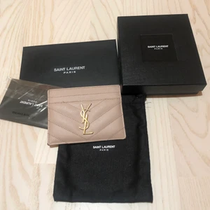YSL korthållare - Korthållare från Yves Saint Laurent - Monogram card case i ”Grain de poudre” i embossed läder.                                           Den är puderrosa/nude med YSL märke i guldtonad metall. 5 stycken kortfack där det får plats 2-3 kort i mittenfacket. Gjord i Italien och köpt i YSL butik i Berlin. Som kan ses på bilder finns slitningar i hörnen och lite guld är bortnött från YSL-märket.                            Kommer med kvitto, äkthetsbevis, kartong och tillhörande dustbag! ✨