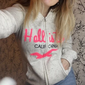 Hollister hoodie  - Vit-grå melerad ’hollister’ hoodie med rosa text och med dragkedja. Hoodien är en lite längre modell och är begagnad men i fint skick. På lappen står det strl XL men passar en M, då den är liten i storleken. Meddela mig vid fler frågor!😊