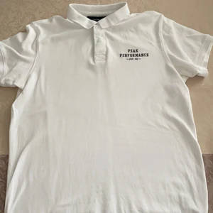 Peak Performance Pikétröja Vit - En vit pikétröja från Peak Performance i storlek M, den är använd och inte i jättebra skick. Ordinarie priset är 699kr från Care of Carl. Det finns trådar som sticker ut vid knapparna, men det syns inte utifrån.
