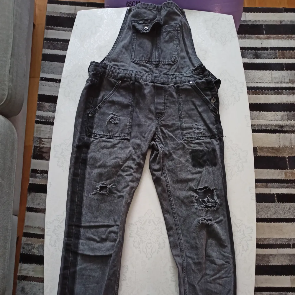 Ett par grå/svarta hängselbyxor i jeansmaterial ifrån Superdry med revor på benen. Byxorna är väl 